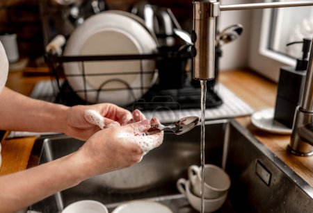 Foto de Girl washing spoon with water at kitchen. Woman cleaning utensil in sink after breakfast during housework - Imagen libre de derechos