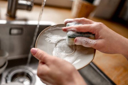 Foto de Girl washing plate dish with sponge and foam at kitchen. Woman cleaning utensil with water in sink during daily rouitine - Imagen libre de derechos
