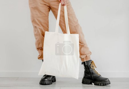 Foto de Girl in boots and pants holding cotton linen eco bag mockup. Woman legs with environment friendly shopping handbag with empty copy space for business - Imagen libre de derechos