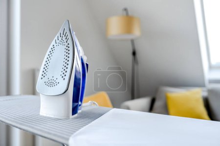 Photo for Electrical iron for wrinkled clothes on ironing board indoor. Modern home appliance tool for housekeeping - Royalty Free Image