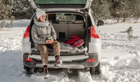 Girl sitting in car trunk in winter time with eco cup mug with hot beverage. Young woman in auto transportation outdoors