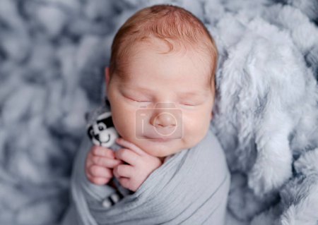 Téléchargez les photos : Mignon bébé garçon nouveau-né emmailloté en tissu gris tenant jouet et dormant sur une couverture en fourrure. Adorable petit enfant enfant sieste portrait studio - en image libre de droit