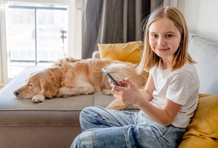 Foto de Preteen girl with golden retriever dog holding smartphone and sitting on sofa at home. Beautiful child kid with labrador doggy pet using cell phone for social media connection - Imagen libre de derechos