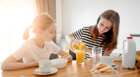 Téléchargez les photos : Girls sisters eat tasty breakfast together and filling glass with orange juice. Female child kid and subling with citrus beverage during morning meal - en image libre de droit