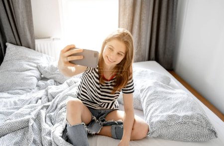 Téléchargez les photos : Pretty girl teenager making selfie in bed with smartphone and smiling. Beautiful young model with mobile cell phone at home - en image libre de droit