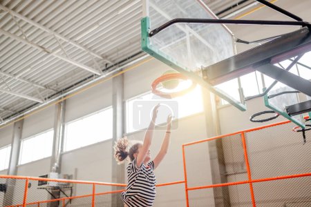 Foto de Pretty girl in trampoline park jumping and holding basketball basket. Happy teenager enjoying amusement activities - Imagen libre de derechos