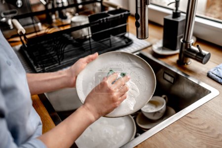 Photo for Girl washing plate dishes at kitchen at home with soap, foam and water. Woman cleaning utensil in sink - Royalty Free Image