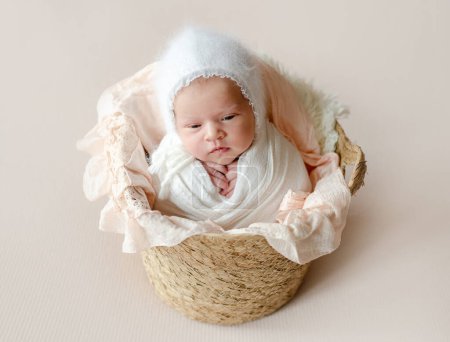 Photo for Newborn baby girl falling asleep lying in basket swaddled in white fabric. Infant child kid wearing knitted hat napping studio portrait - Royalty Free Image