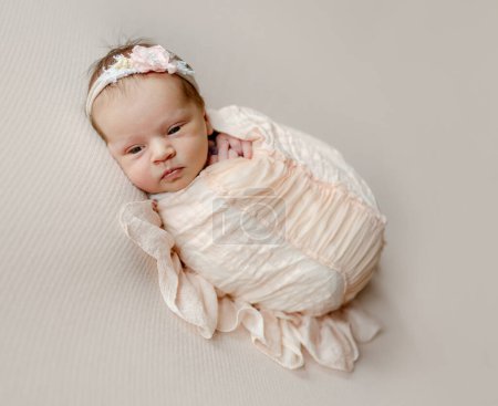 Téléchargez les photos : Newborn baby girl wearing flower wreath falling asleep. Infant child kid lying swaddled in peach fabric studio portrait - en image libre de droit