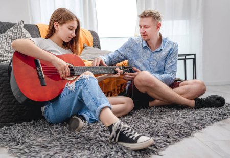 Photo for Father guy man teaching girl teenager daughter guitar playing at home sitting on floor. Family musical lessons with strings instrument - Royalty Free Image