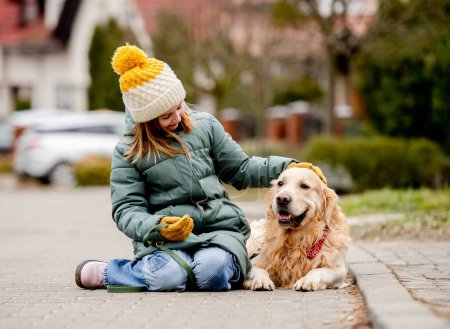 Foto de Niña preadolescente sentada con perro golden retriever en asfalto en la calle de otoño con sombrero y chaqueta caliente. Niño bonito acariciando pura raza mascota perrito labrador al aire libre en la ciudad - Imagen libre de derechos