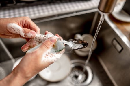 Foto de Girl washing spoon with water at kitchen. Woman cleaning utensil in sink after breakfast during housework - Imagen libre de derechos