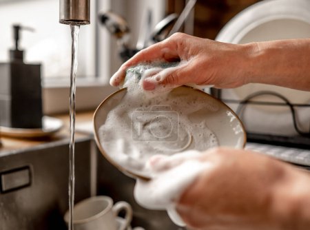 Photo for Girl washing plate dish with sponge and foam at kitchen. Woman cleaning utensil with water in sink during daily rouitine - Royalty Free Image