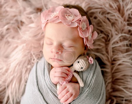 Foto de Bebé recién nacido niña durmiendo sosteniendo muñeca de punto princesa juguete y usando corona de flores. Niño lindo niño siesta en piel envuelta en tela - Imagen libre de derechos