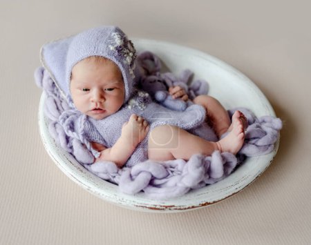 Foto de Bebé recién nacido niña con sombrero de punto y traje sosteniendo dulce juguete de conejito. Niño pequeño acostado en lavabo color púrpura estudio retrato - Imagen libre de derechos