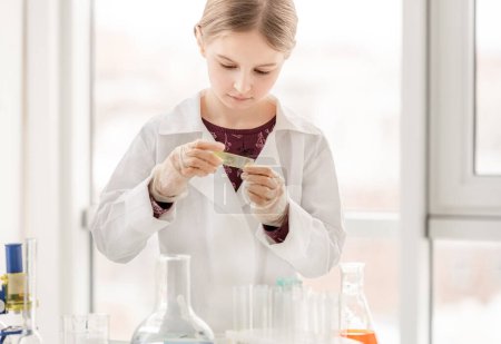 Foto de Chica inteligente durante el experimento de química científica mirando el vidrio con pruebas. Colegiala con equipo químico en la lección escolar - Imagen libre de derechos