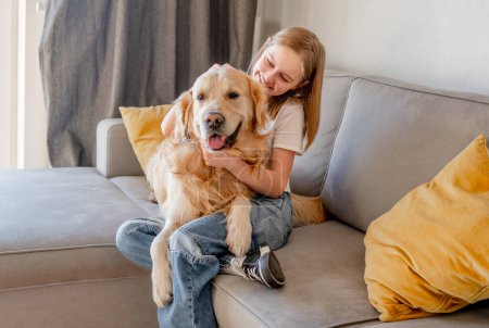 Photo for Preteen girl hugging golden retriever dog and smiling sitting on sofa at home. Beautiful child kid with labrador doggy pet indoors - Royalty Free Image