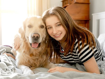 Téléchargez les photos : Pretty girl hugging golden retriever dog and smiling sitting in bed. Female teenager with purebred doggy pet labrador looking at camera at home - en image libre de droit