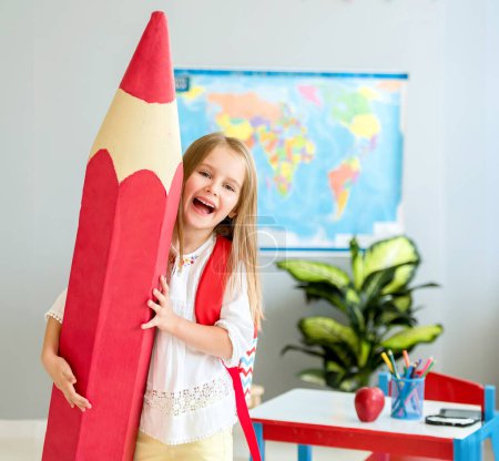 Photo for Little smiling blond girl with red bag holding huge red decorative pencil on her shoulder in the school classroom - Royalty Free Image
