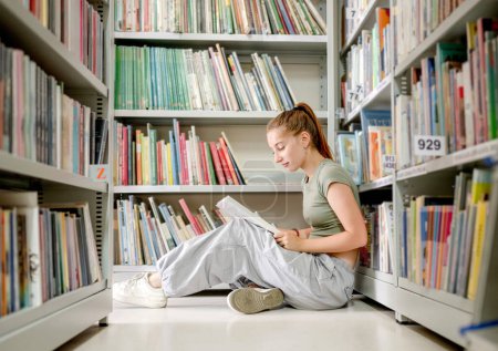 Photo for Girl teenager sitting in library on floor and reading book indoors. Beautiful student preparing for exam with education mterials near bookshelfs - Royalty Free Image