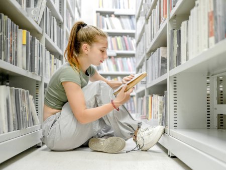 Photo for Teenager girl sitting in library on floor and reading academic book. Beautiful female student preparing for exam with education mterials in bookstore - Royalty Free Image