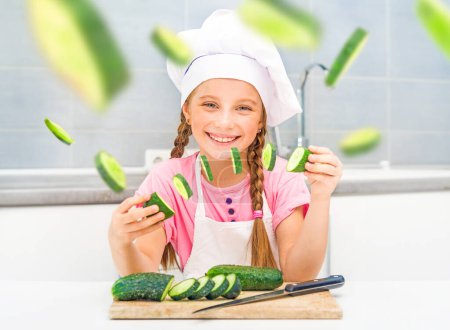 Foto de Sonriente niña corta pepinos a casa en la cocina - Imagen libre de derechos