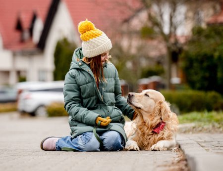 Photo for Preteen child girl sitting with golden retriever dog on asphalt at autumn street wearing hat and warm jacket. Pretty kid petting purebred pet doggy labrador outdoors at city - Royalty Free Image