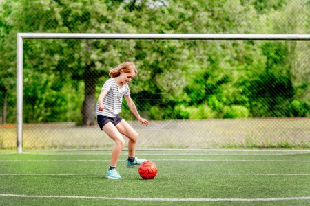 Foto de Niña bonita corriendo con pelota de fútbol en la hierba durante el juego. Lindo niño femenino en el campo de fútbol jugando juego activo - Imagen libre de derechos