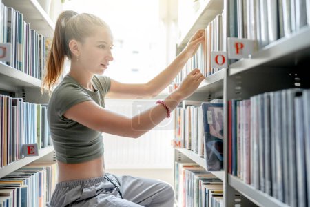 Foto de Pretty girl teenager in library looking for book at bookshelf indoors. Beautiful female student in casual clothes searching for literature - Imagen libre de derechos