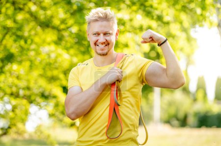 Photo for Man with elastic rubber band showing muscles outdoors after arm workout. Guy with sport equipment looking at camera and smiling - Royalty Free Image