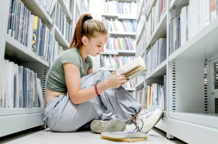 Téléchargez les photos : Adolescente assise à la bibliothèque sur le sol et lisant un livre académique. Belle étudiante se préparant à l'examen avec des articles d'éducation en librairie - en image libre de droit