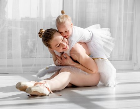 Foto de Joven madre en vestido de ballet cisne abrazando a la pequeña hija sonriente sentada en el suelo en la sala de luz. Emociones positivas de madre e hija en el estudio de ballet - Imagen libre de derechos