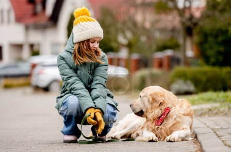 Téléchargez les photos : Preteen enfant fille assis avec chien golden retriever à la rue automne portant un chapeau et une veste chaude. Joli gosse avec chien de race labrador en plein air à la ville - en image libre de droit