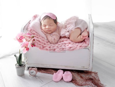 Téléchargez les photos : Adorable petite fille nouveau-née dormant dans un petit lit sur une couverture tricotée. Enfant mignon enfant en robe rose sieste avec décoration de fleurs d'orchidée - en image libre de droit