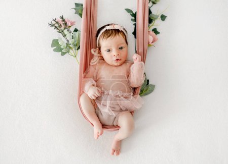 Entzückendes neugeborenes Mädchen, das in der Hängematte sitzt und einen Kranz trägt. Niedliches Kleinkind in Schaukel mit Blumendekor