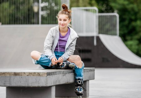 Foto de Cute girl roller skater sitting in city park and smiling. Pretty female teenager posing during rollerskating - Imagen libre de derechos
