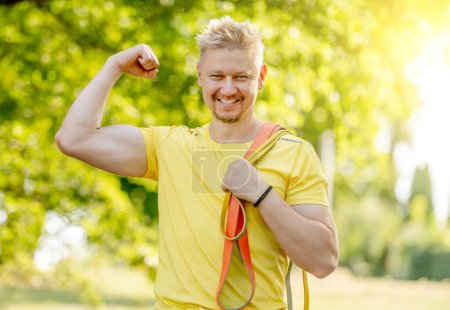 Foto de Hombre con goma elástica mostrando los músculos al aire libre después del entrenamiento del brazo. Chico con equipo deportivo mirando a la cámara y sonriendo - Imagen libre de derechos