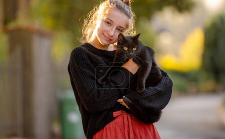 Photo for Pretty girl hugging black cat outdoors at street with autumn light. Beautiful model teenager holding feline animal at park - Royalty Free Image