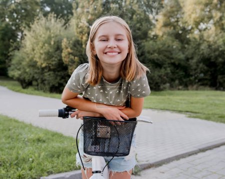 Téléchargez les photos : Jolie fille préadolescente avec vélo à l'extérieur regardant la caméra et souriant. Jolie enfant avec vélo dans la rue de la ville au parc d'été - en image libre de droit