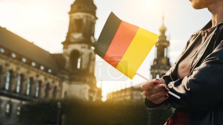 Téléchargez les photos : Jeune femme tient le drapeau allemand dans la main, avec fond de ville floue en automne - en image libre de droit