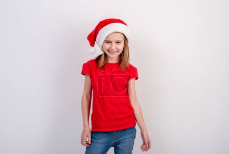 Foto de Niña en camiseta roja y sombrero de Santa posa para una foto festiva sobre un fondo blanco - Imagen libre de derechos