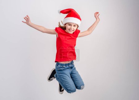 Foto de Niña con camiseta roja y sombrero de Santa está saltando en una foto festiva sobre un fondo blanco - Imagen libre de derechos