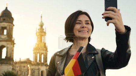 Foto de Joven mujer toma selfie con bandera alemana en la mano, en contra de la ciudad borrosa telón de fondo en otoño - Imagen libre de derechos