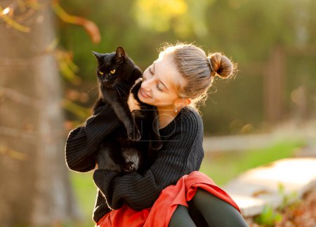 Photo for Pretty girl in red skirt hugging black cat outdoors at street with autumn leaves. Beautiful model teenager sitting with feline animal at park - Royalty Free Image