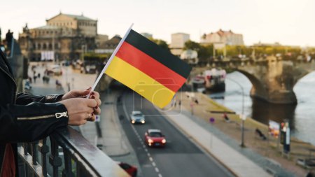Foto de Mujer joven sostiene bandera alemana en la mano, con fondo borroso de la ciudad en otoño - Imagen libre de derechos
