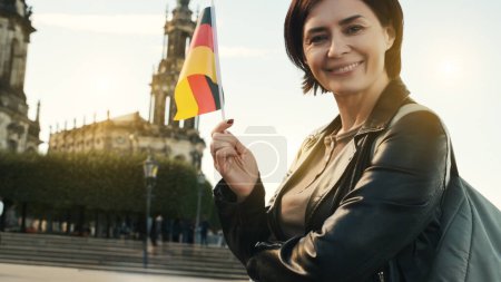 Foto de Mujer joven sonríe con la bandera alemana en la mano, contra el fondo borroso de la ciudad en otoño - Imagen libre de derechos