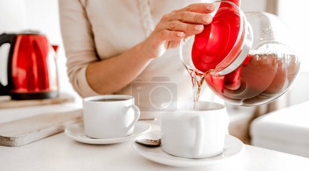 Photo for Girl pouring with hot tea from teapot cups at kitchen. Woman preparing warm herbal beverage with kettle and mugs - Royalty Free Image