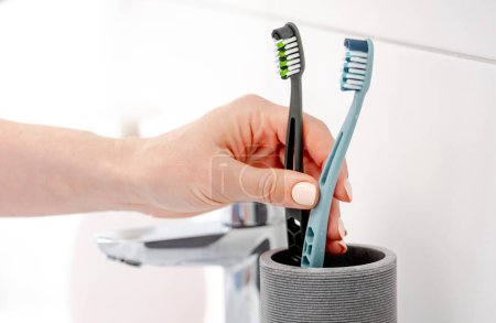 Photo for GirlS Hand Places Two Toothbrushes In Cup For Hygiene In Bright Bathroom - Royalty Free Image