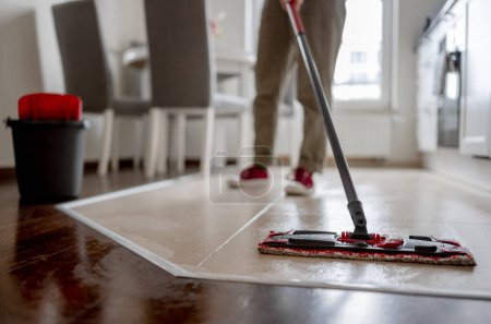 Photo for Woman Mops Floor In Kitchen With Broom Prominently In Foreground - Royalty Free Image