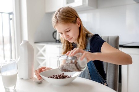 Photo for Sweet Girl Pours Flakes Into Plate For Dry Breakfast In Kitchen - Royalty Free Image